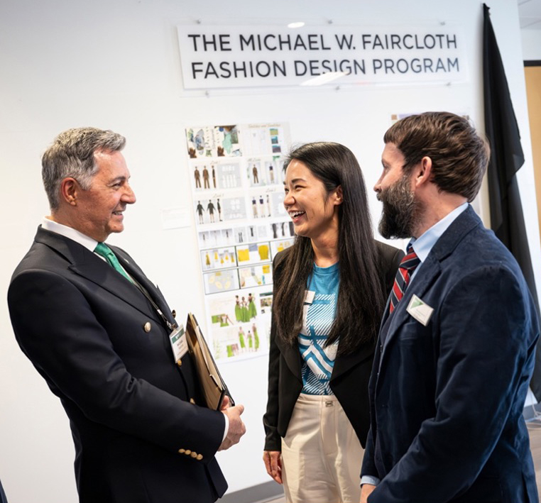 Three people, Michael Faircloth, Chanjuan Chen and Jeremy Bernardino, talking under the Michael W. Faircloth Fashion Design Program sign.