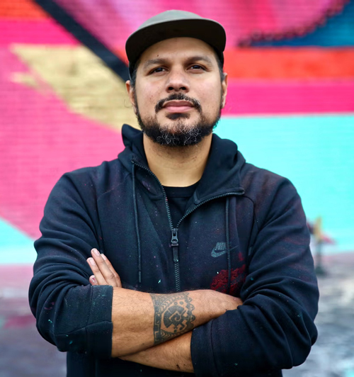 Victor Quinonez is standing with his arms crossed. He wears a dark jacket and a baseball cap.