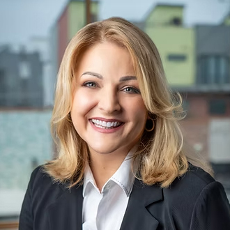Bonnie is facing forward and smiing. She has blond wavy hair and wears a blue jacket over a white shirt.