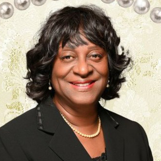 Carolyn is facing forward and smiling. She has dark curly hair and wears a dark suit.