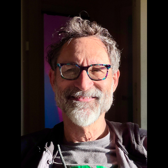 Hepi is facing forward and smiling. He has short hair a white beared and wears glasses, a white shirt and tie with a turquoise-blue jacket.