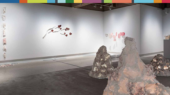 Gallery corner with a vertical hanging of small ceramic wall pieces, rough pink and tan ceramic peaks rising from the floor. 
